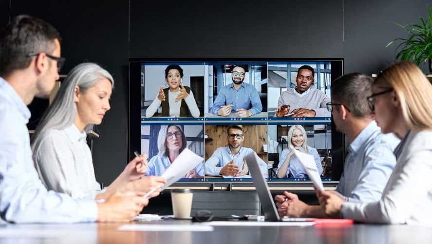 people are sitting at a table with laptops and a tv with a zoom meeting