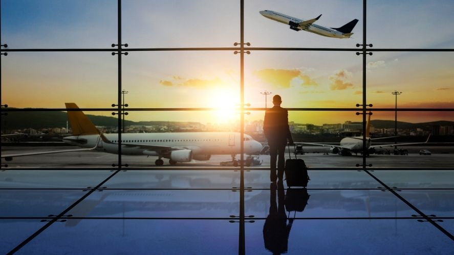 Business traveler standing in airport with luggage, looking out windows at airplanes taking off